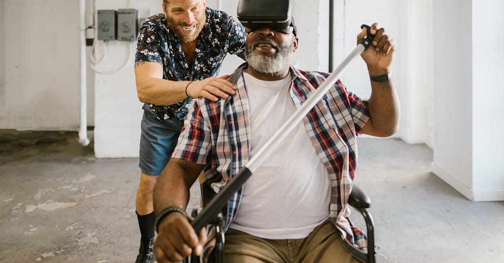 Man assists wheelchair user engaged in rehabilitation exercise with VR set indoors.